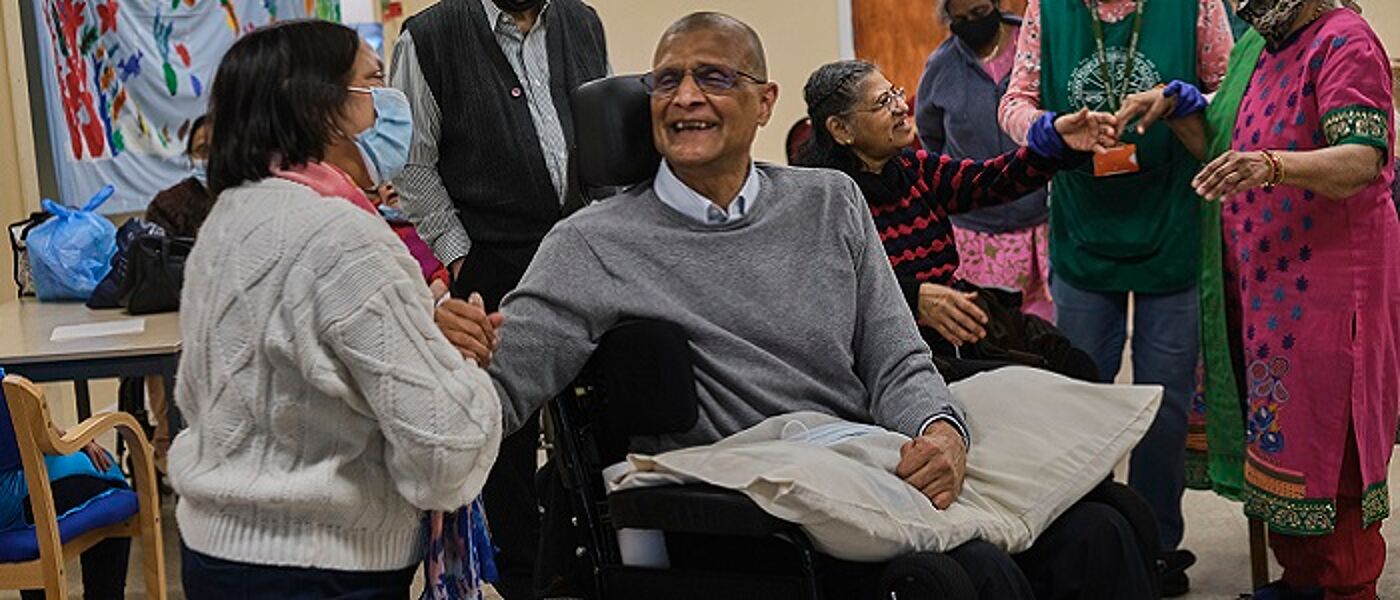 In a community room, an older Asian man in a wheelchair holding hands and smiling at a woman. There are other people chatting in the background. Source: Centre for Aging Better https://ageingbetter.resourcespace.com/pages/view.php?ref=11876&k=&search=age%2C+positive%2C+image%2C+library%2C+%40%40339&offset=0&order_by=resourceid&sort=DESC&archive=