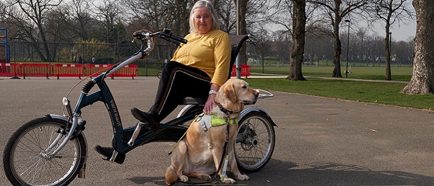 A middle-aged woman using a tricyle in a park with her guidedog siting alongside her. Source: Centre for Better Aging https://ageingbetter.resourcespace.com/pages/view.php?ref=11437&k=&search=11883%2C+%40%40339&offset=0&order_by=&sort=DESC&archive=