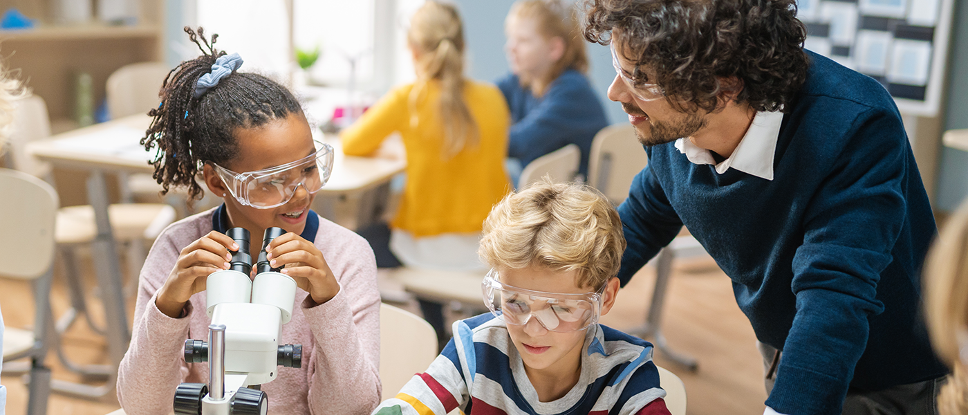 Two children are sat in a classroom wearing safety goggles. One is looking into a microscope. They are supervised by a man and are smiling.