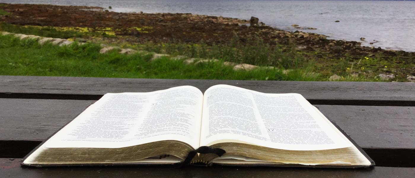 A large bible lying open on top of a wooden picknick table with  the shore of a Scottish lock in the background