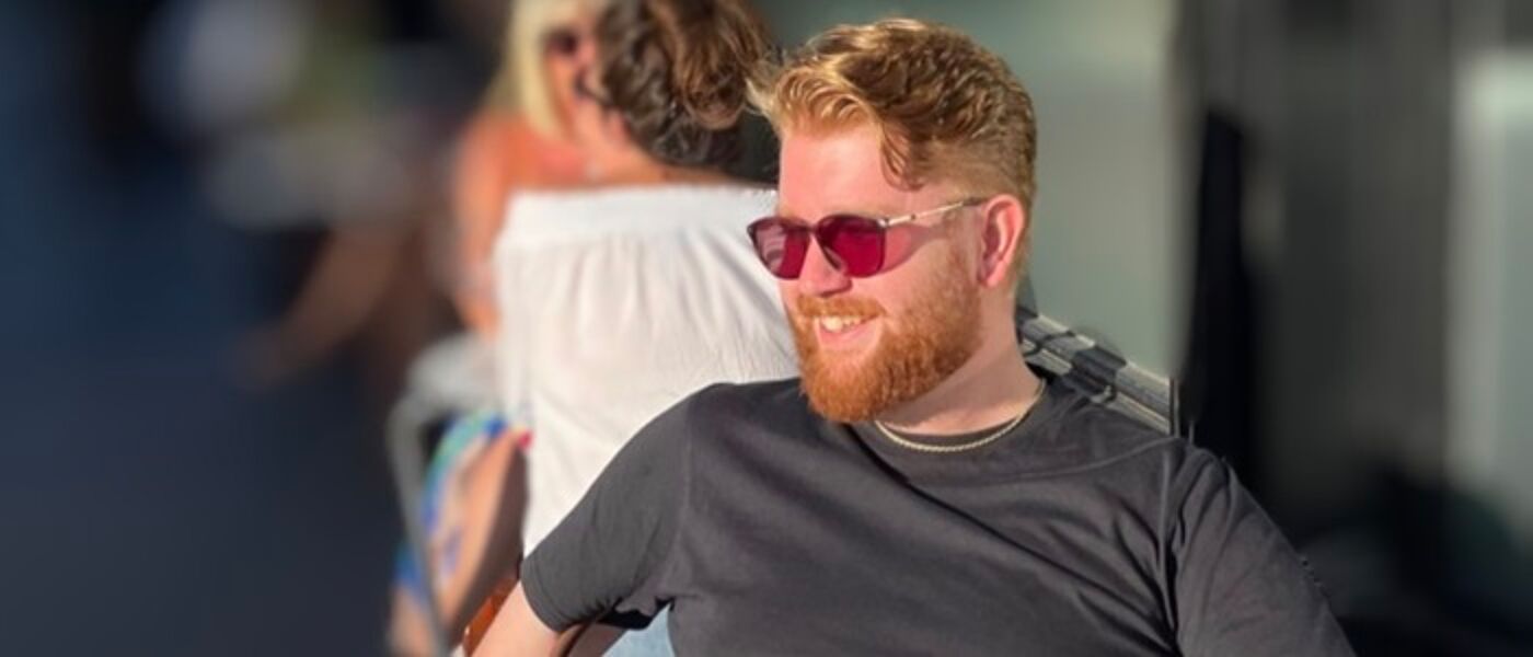 Man with red hair, a beard and sunglasses wearing a grey t-shirt looking and smiling to the side