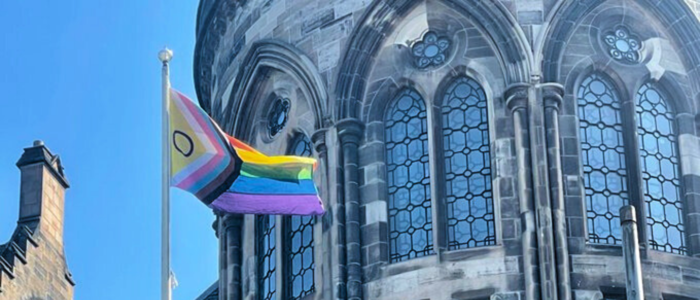 A photo of the Progress Pride flag flying in the wind on the top of a flag pole outside the University of Glasgow Main Building