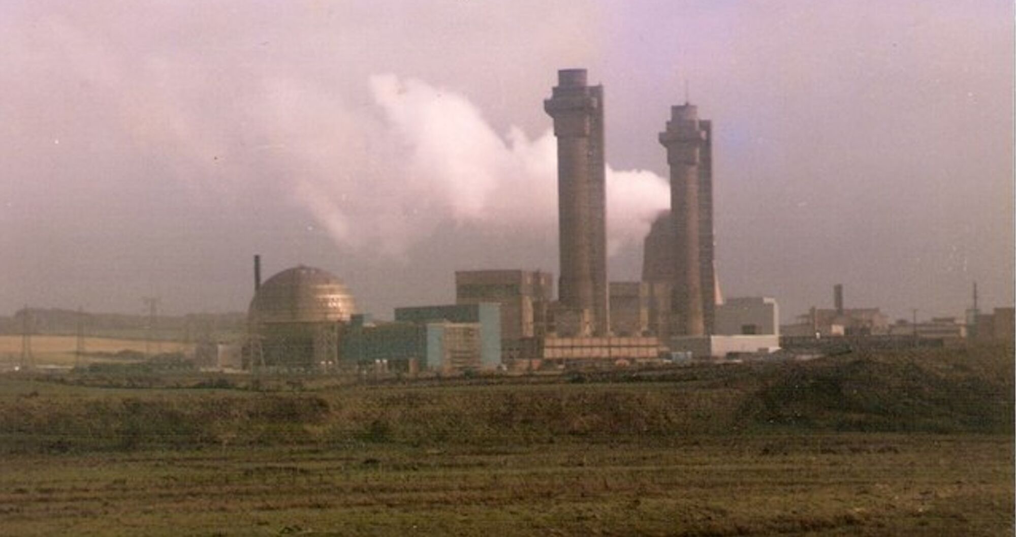 A view of the buildings of Sellafield nuclear power station with steam plumes emitting from it, seen across a field and against a stormy sky