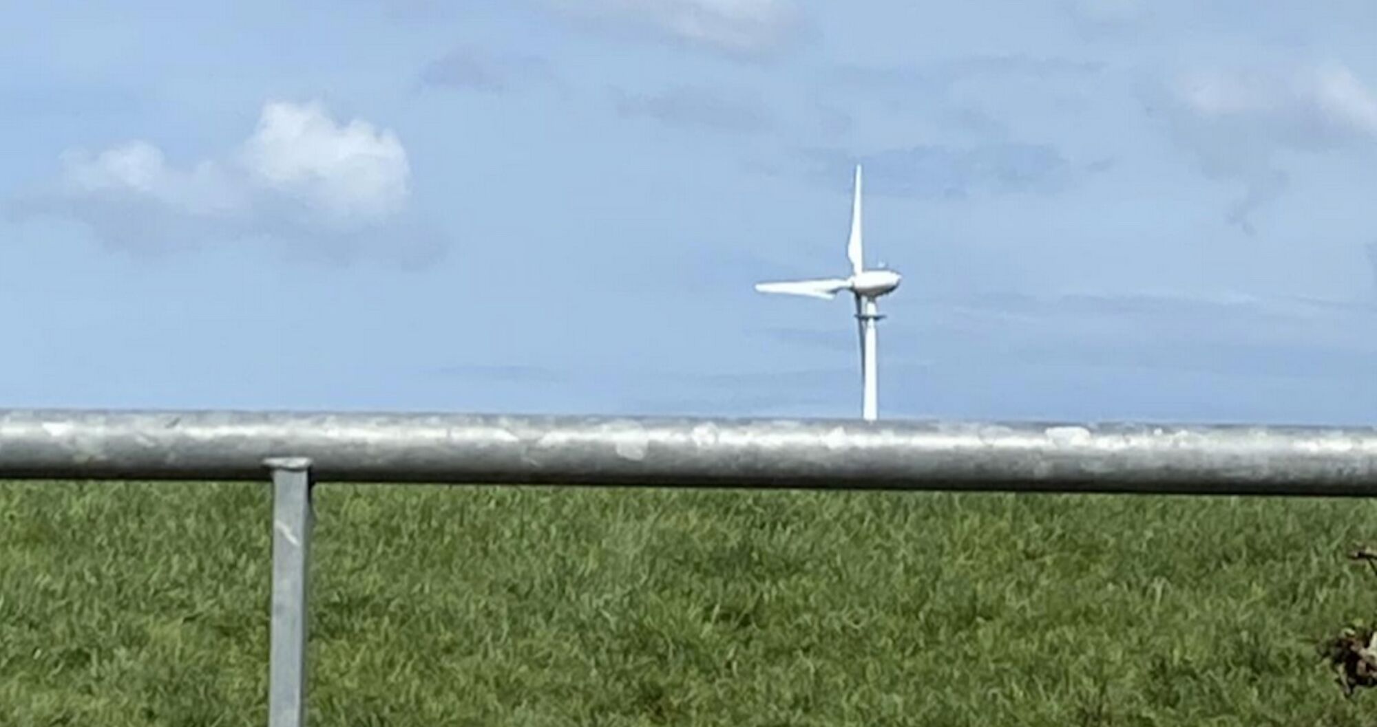 A wind turbine in a field beyond a metal gate, seen against a blue sky