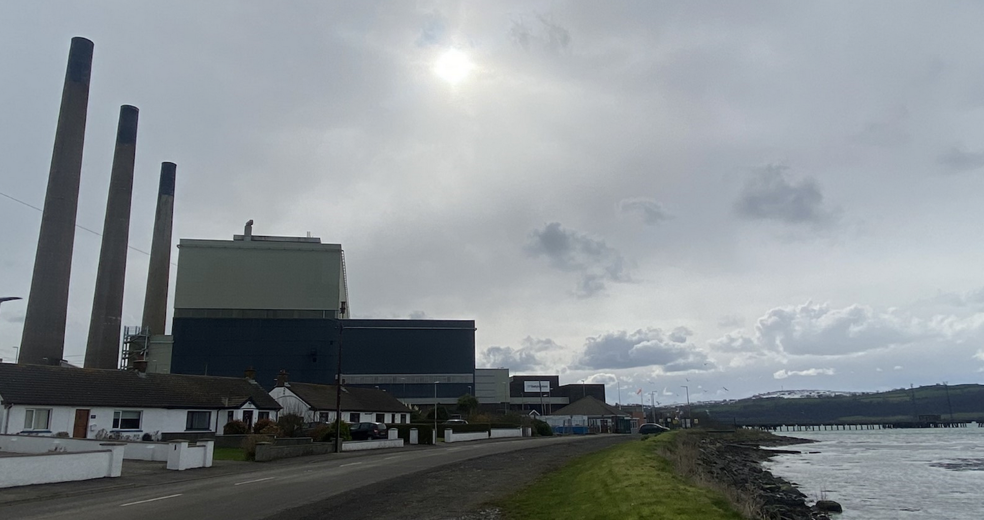 A power station with three tall thin chimneys and a square building, with small white cottages and a bay of the sea in front of it. Photo credit: Ewan Gibbs