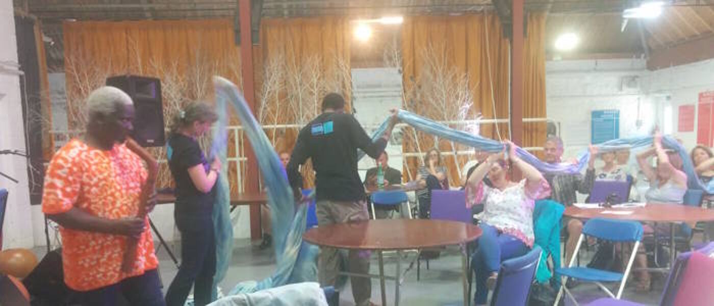 Alison Phipps and Tawona Sitholé passing a long bolt of blue cloth throughout a seated crowd while Gameli Tordzro plays a wind instrument