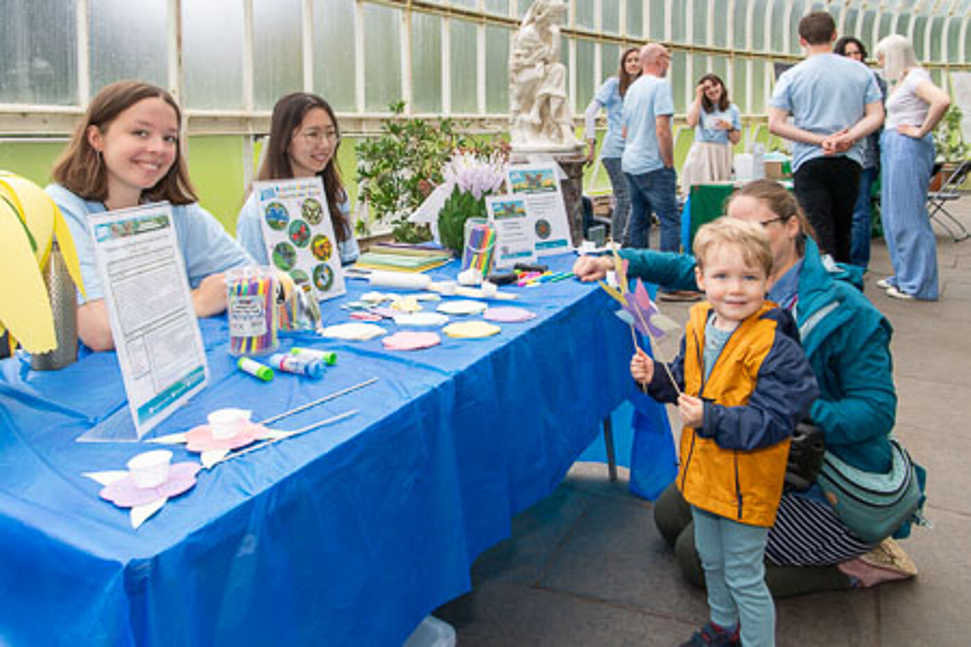 Two GSF volunteers sitting behind a table with craft materials and blue table cloths. A child is standing holding a paper flower and smiling at the camera.