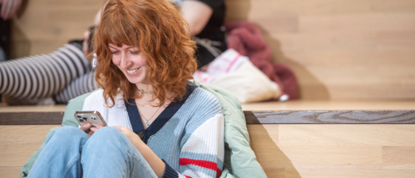 A student sitting in the James McCune Smith Building looking and smiling at their phone