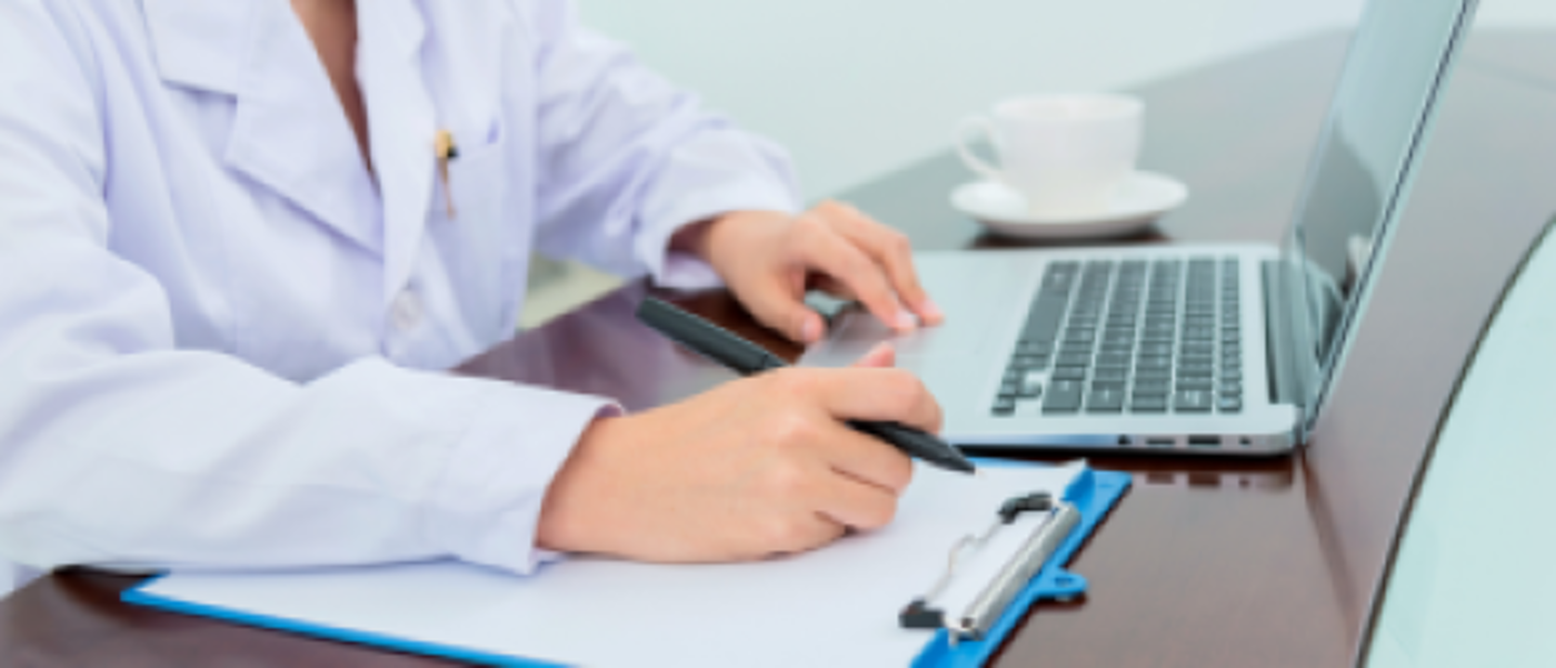 Medical practitioner reviewing notes at a desk using both a laptop and paper form on a clipboard