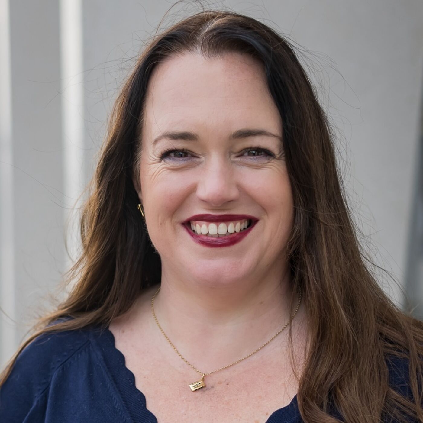 Person wearing a navy blue top with a scalloped neckline and a delicate gold necklace featuring a small rectangular pendant. The background is a plain light gray wall