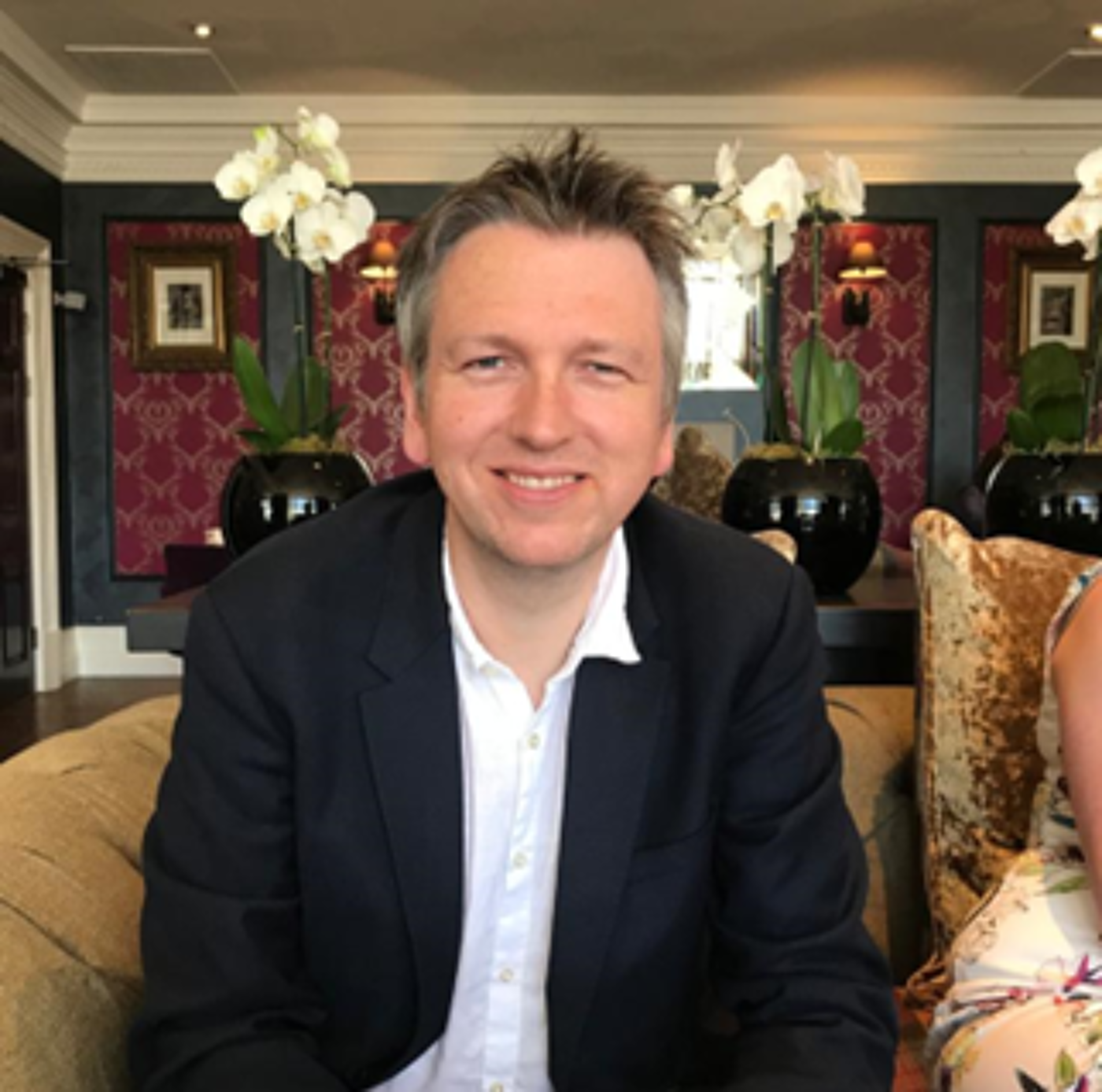 Person wearing a dark suit jacket over a white shirt, seated on a beige sofa in an elegant room. The background features decorative wallpaper with a red and gold pattern, framed artwork, and large black vases holding white orchids.