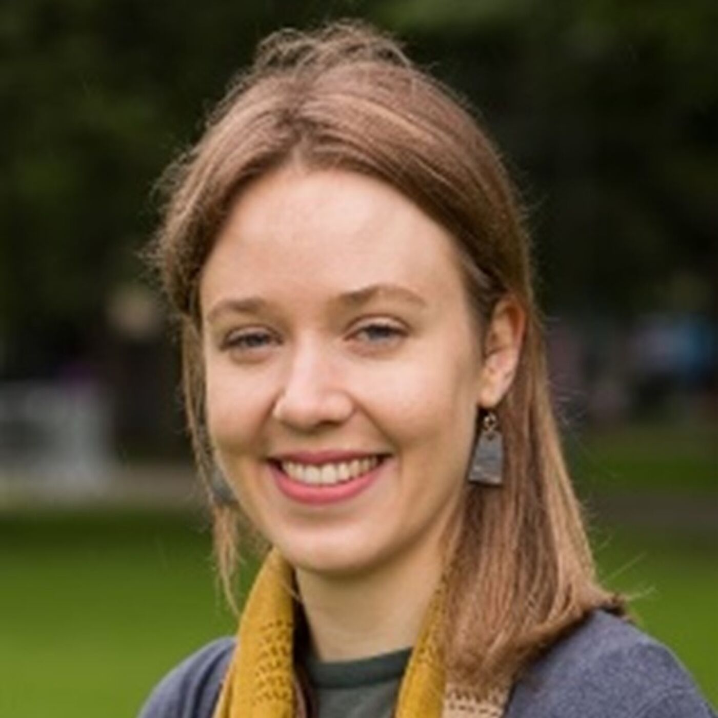 Person with shoulder-length light brown hair wearing a mustard yellow scarf and dark top, standing outdoors on a grassy area with blurred trees in the background.
