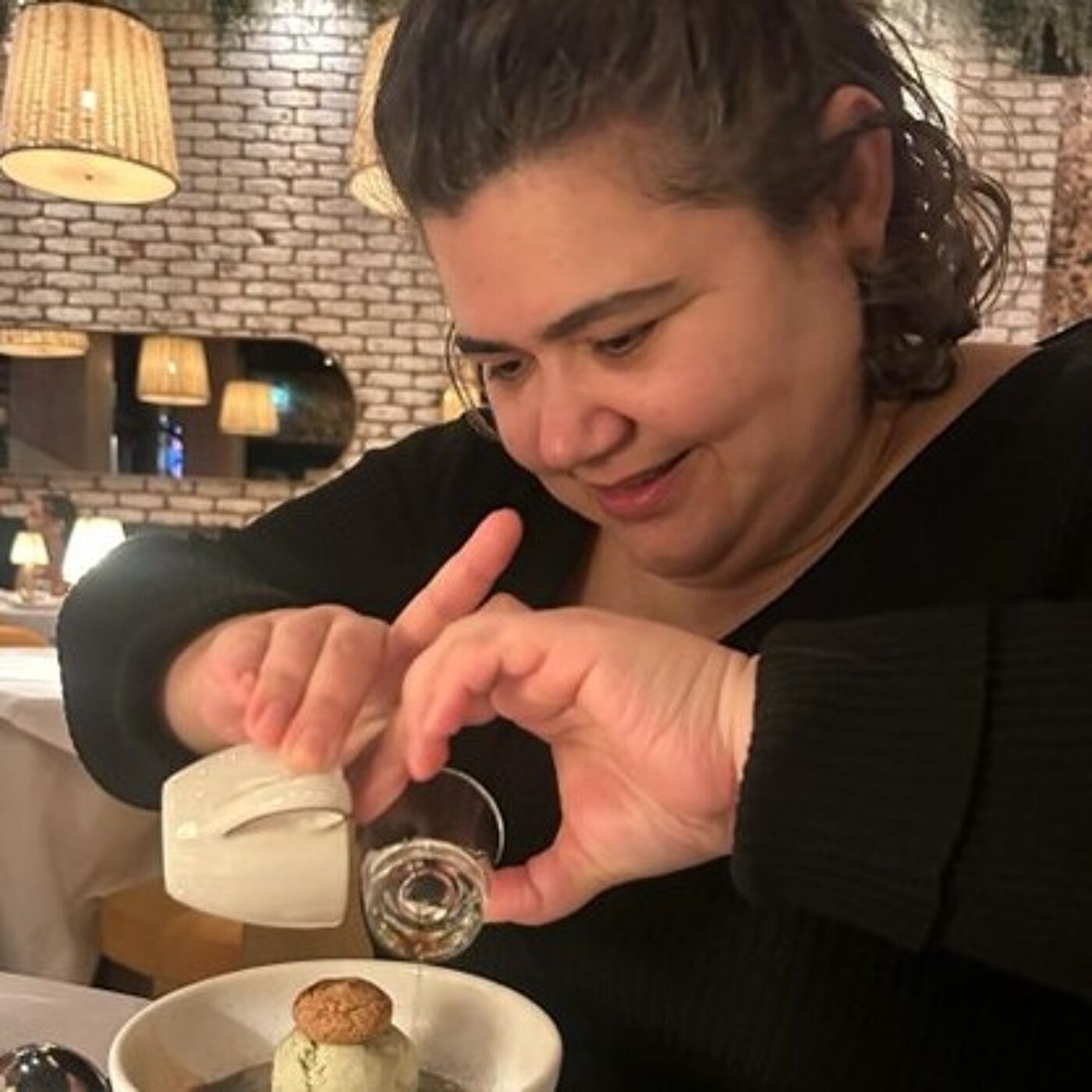 Person seated at a restaurant table, pouring a clear liquid from a small glass into a dessert bowl that contains a scoop of ice cream topped with a biscuit. The setting features warm ambient lighting, brick walls, and large hanging lamps.