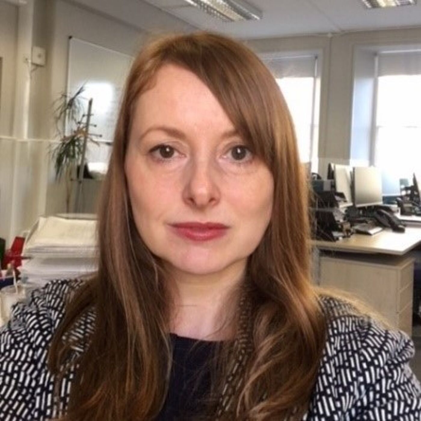 Person with long, light brown/red hair wearing a patterned jacket over a dark top, standing in an office environment. The background shows desks with computer monitors, stacks of papers, a whiteboard, and large windows letting in natural light