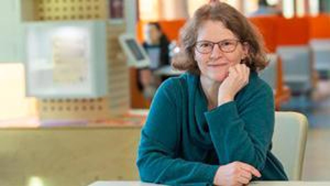 Prof Petra Meier sitting in study area facing camera with hand on chin and chairs and students in background