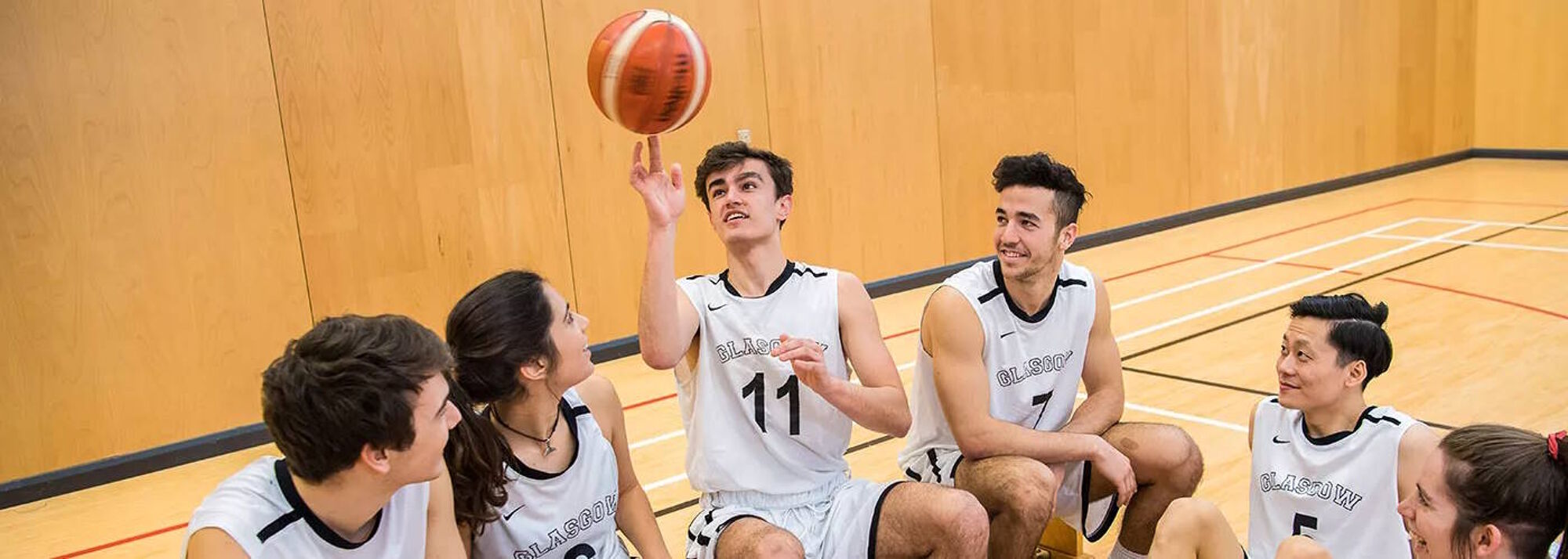 Six basketball players sitting on a bench in their white team kit. The middle player is holding a ball in the air.