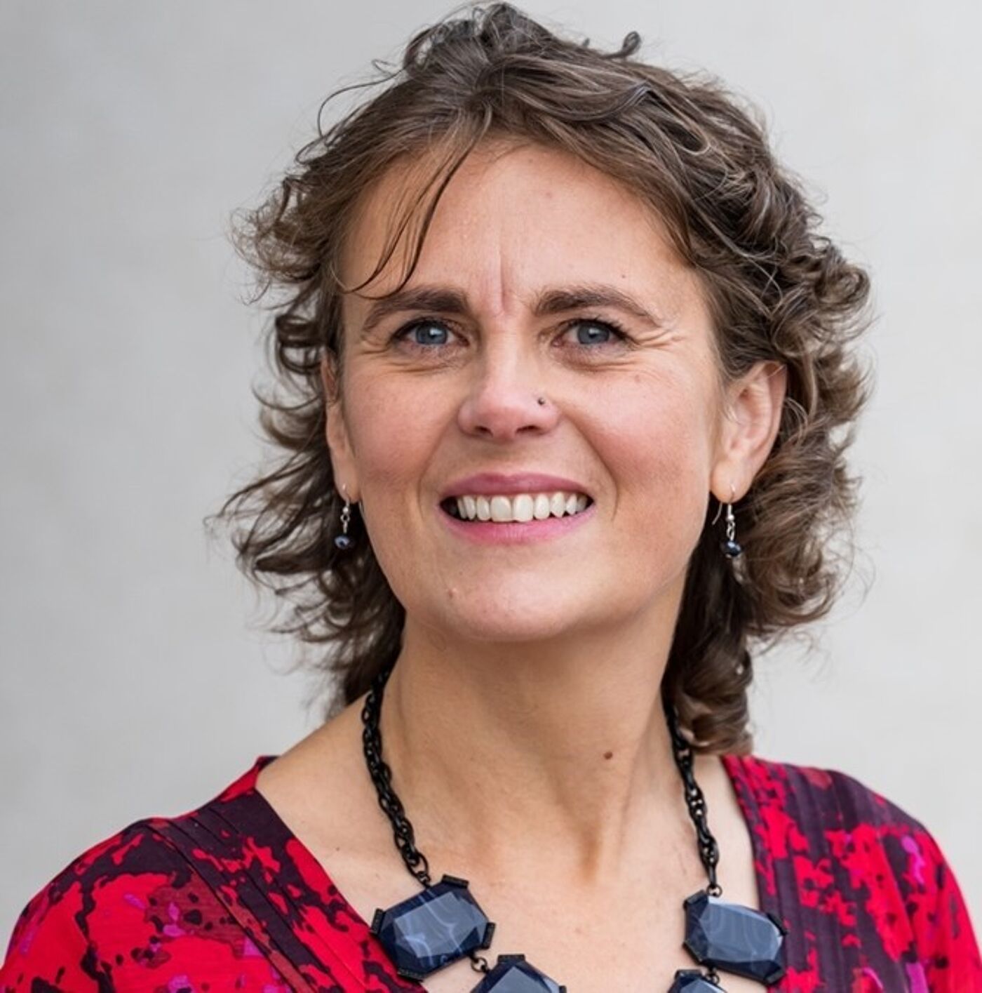 Person wearing a red and black patterned top and a chunky black geometric necklace, with short curly hair. The background is a plain light gray wall.