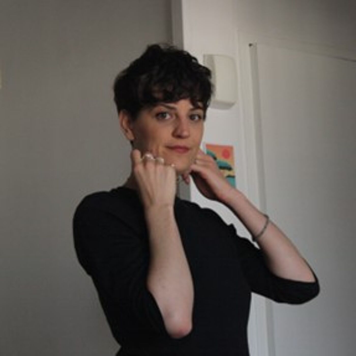 Person wearing a black long-sleeve top, standing indoors near a white wall and door. A small colorful artwork is partially visible on the wall behind.