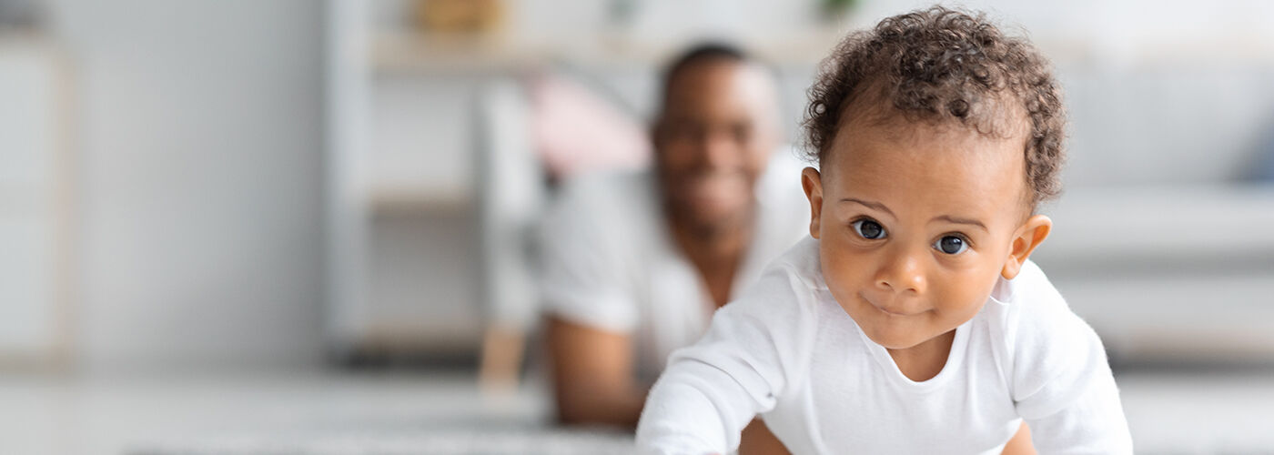 A baby is crawling towards the camera with dad lying on the floor behind