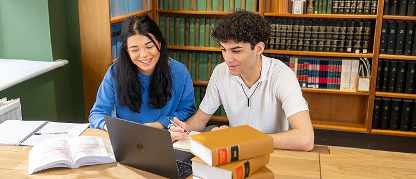 An image of two students working together at a desk with a laptop on the table, and bookshelves behind them.