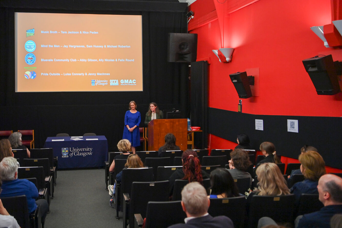 two people presenting to a group in the Andrew Stewart Cinema. text projected behind them displays the names of students and organisations taking part in the Applied Dissertation