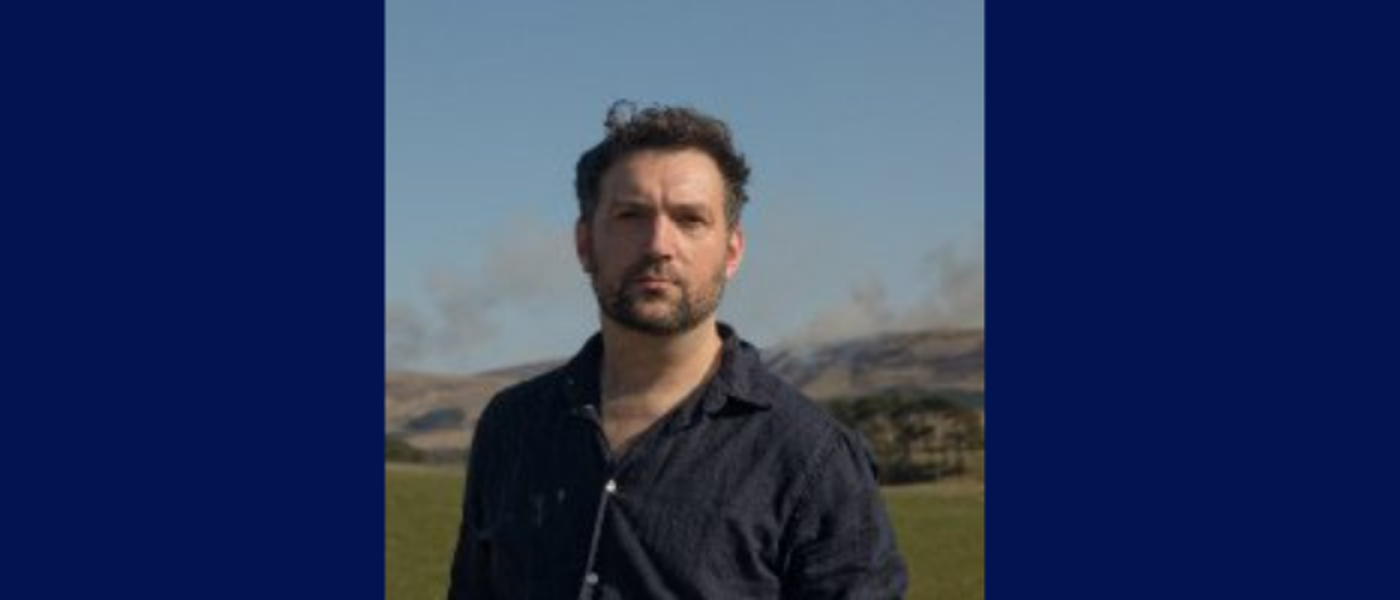 Man in dark shirt standing in front of a scenic background on a dark blue background.