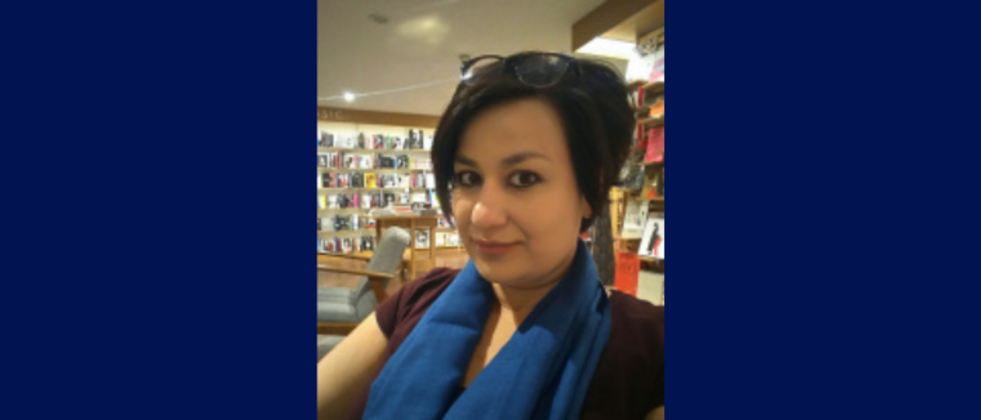 Woman in a bookshop with glasses on her head, wearing a blue scarf and brown t-shirt on a blue background.