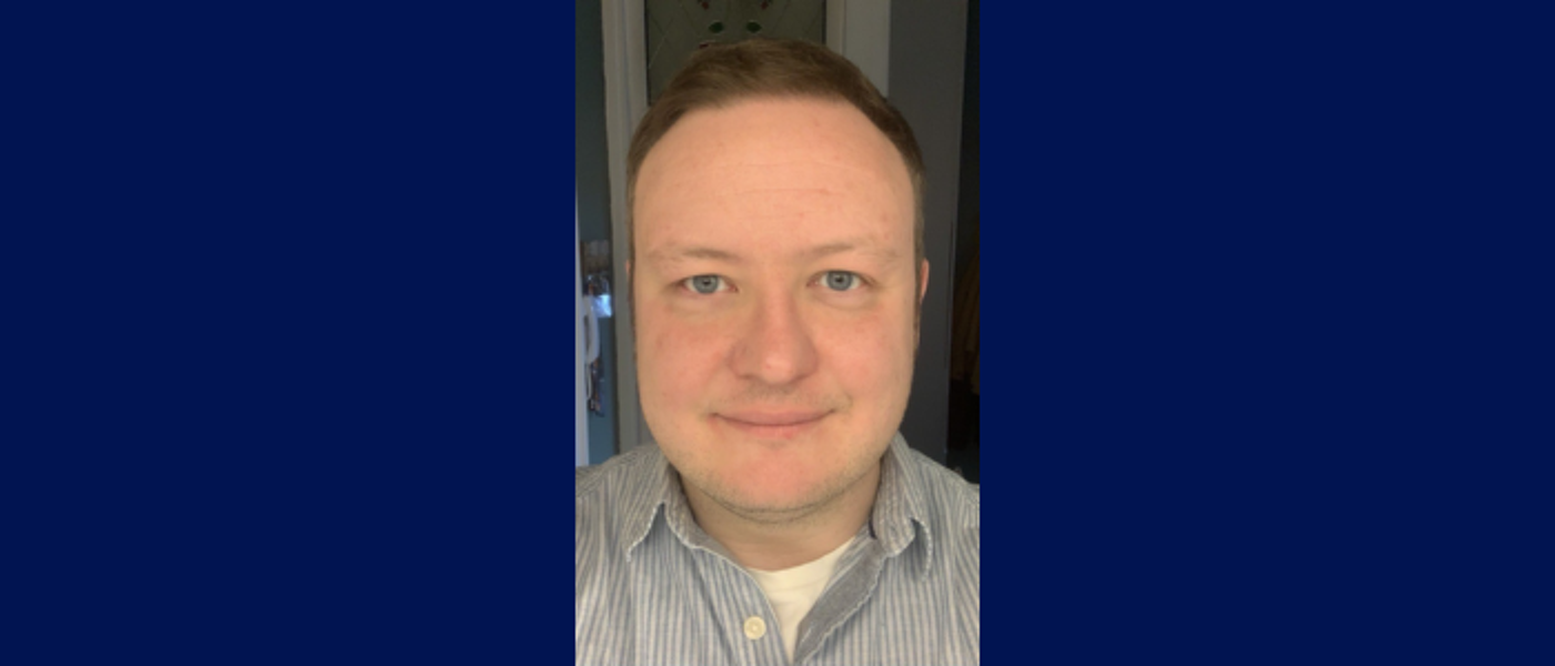 Man looking directly at the camera in a light blue striped shirt. On a dark blue background.