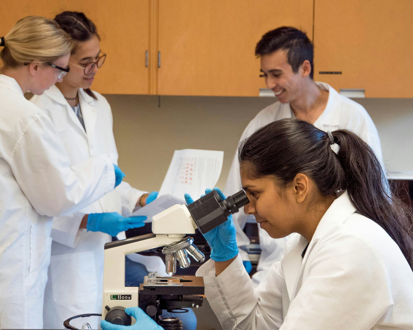 This picture depicts for scientists in lab coats conducting research. To the left, two women look over a paper together. In the right hand background a man is smiling. In the foreground a woman looks through a microscope