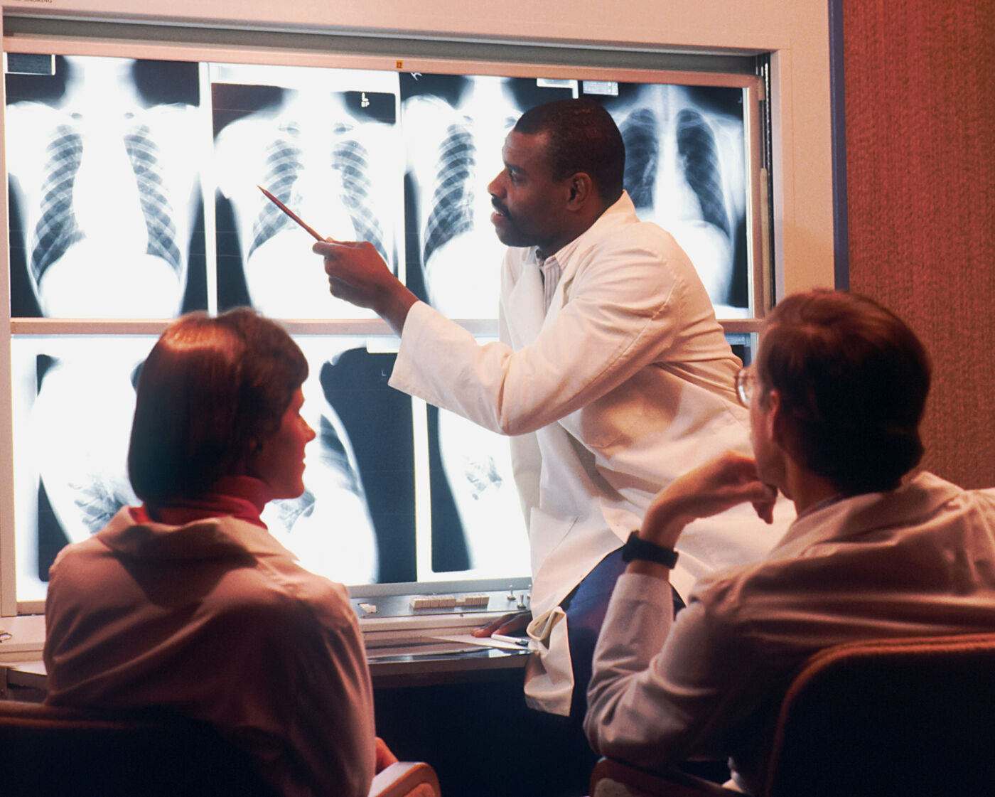 Chest X-rays are projected onto the wall behind three scientists. One scientist is standing and inspecting the x rays, while the other two sit and watch