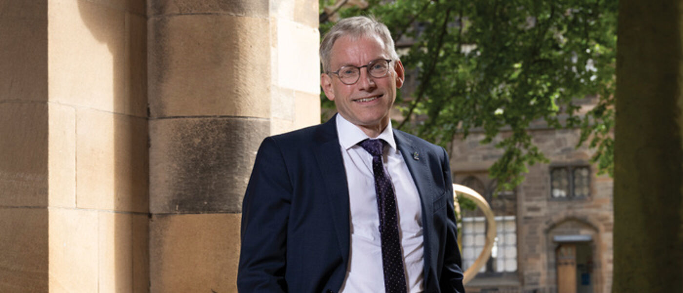 Prof Andy Schofield standing in the cloisters wearing a navy suit and smiling to the camera