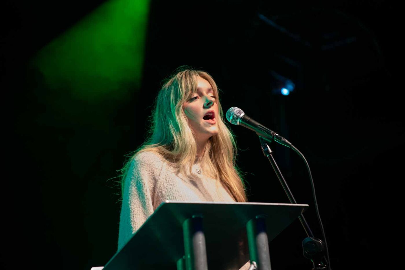 A young woman with blonde hair singing on a stage, only her head and shoulders are in frame