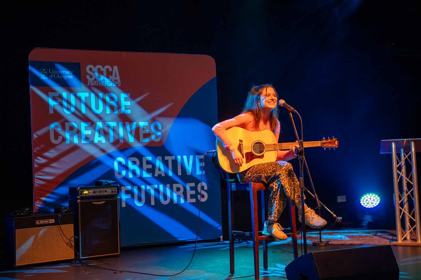 a young woman singing with a guitar on stage with a banner that says ' FUTURE CREATIVES CREATIVE FUTURES ' on it in the background