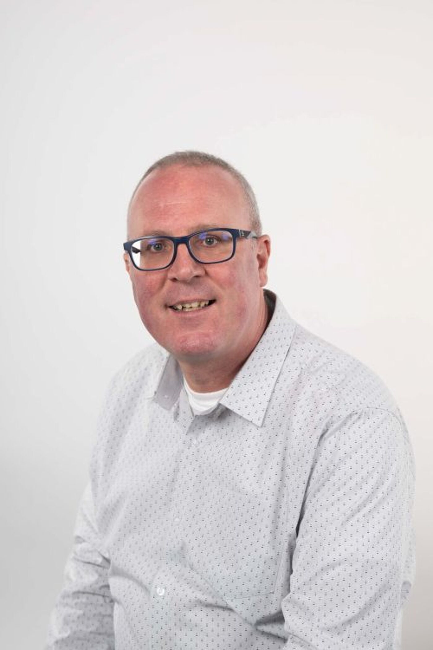 A person wearing a plain white button-up shirt is seated against a simple, light-colored background. The shirt has a collar and appears neatly pressed, giving a clean and professional appearance.