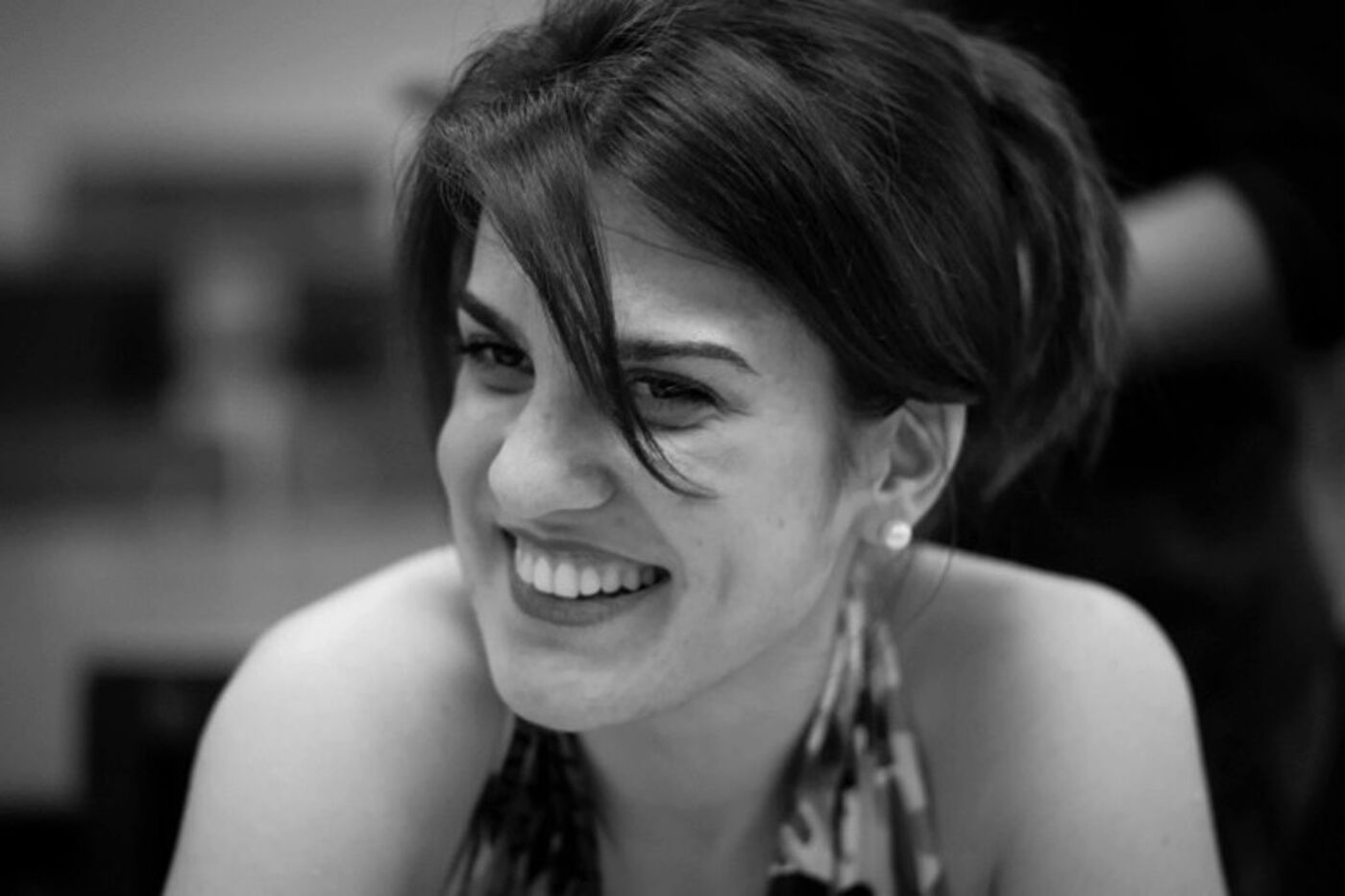 A black-and-white photo of a person with short, styled hair wearing a patterned halter-neck top. The individual is leaning slightly forward, and the background appears softly blurred, suggesting an indoor setting.