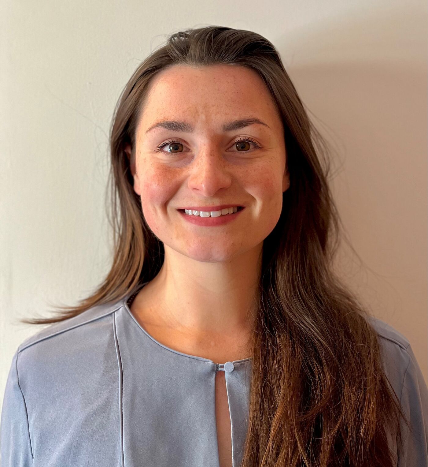 A person with long, wavy brown hair is wearing a light blue top with a round neckline. The background is a plain, light-colored wall, creating a simple and neutral setting.