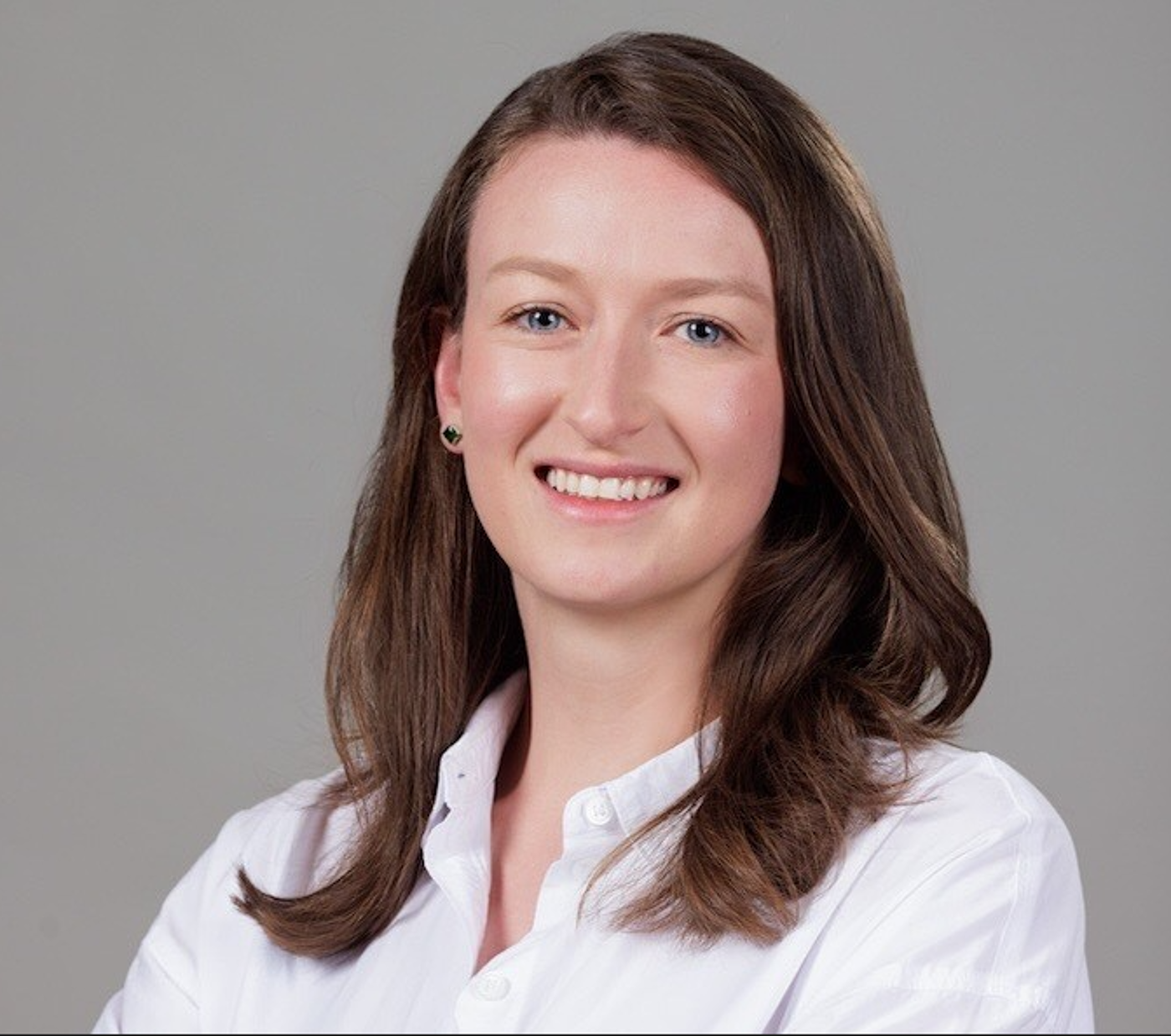 A person with shoulder-length brown hair is wearing a white button-up shirt and is posed against a plain gray background. The arms are crossed in front, giving a professional and confident appearance.