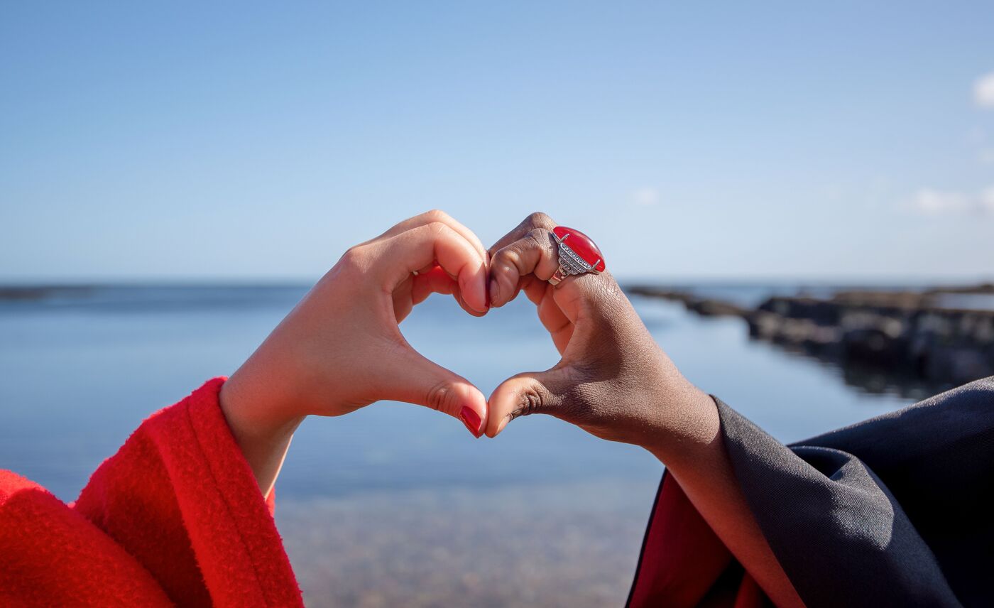 Two hands forming the shape of a heart against a backdrop of a calm body of water and a clear blue sky. One hand is wearing a bright red sleeve and a ring with a large red stone, while the other hand is in a dark sleeve. The horizon and rocky shoreline are visible in the distance.