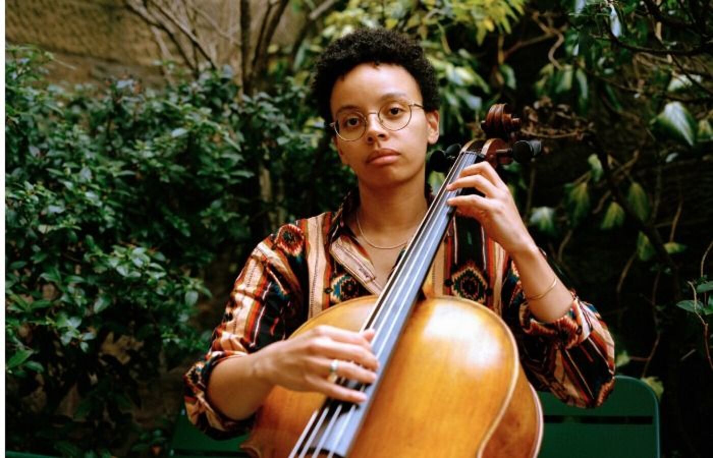 Simone Seales sits holding a cello, wearing a striped shirt and with trees behind
