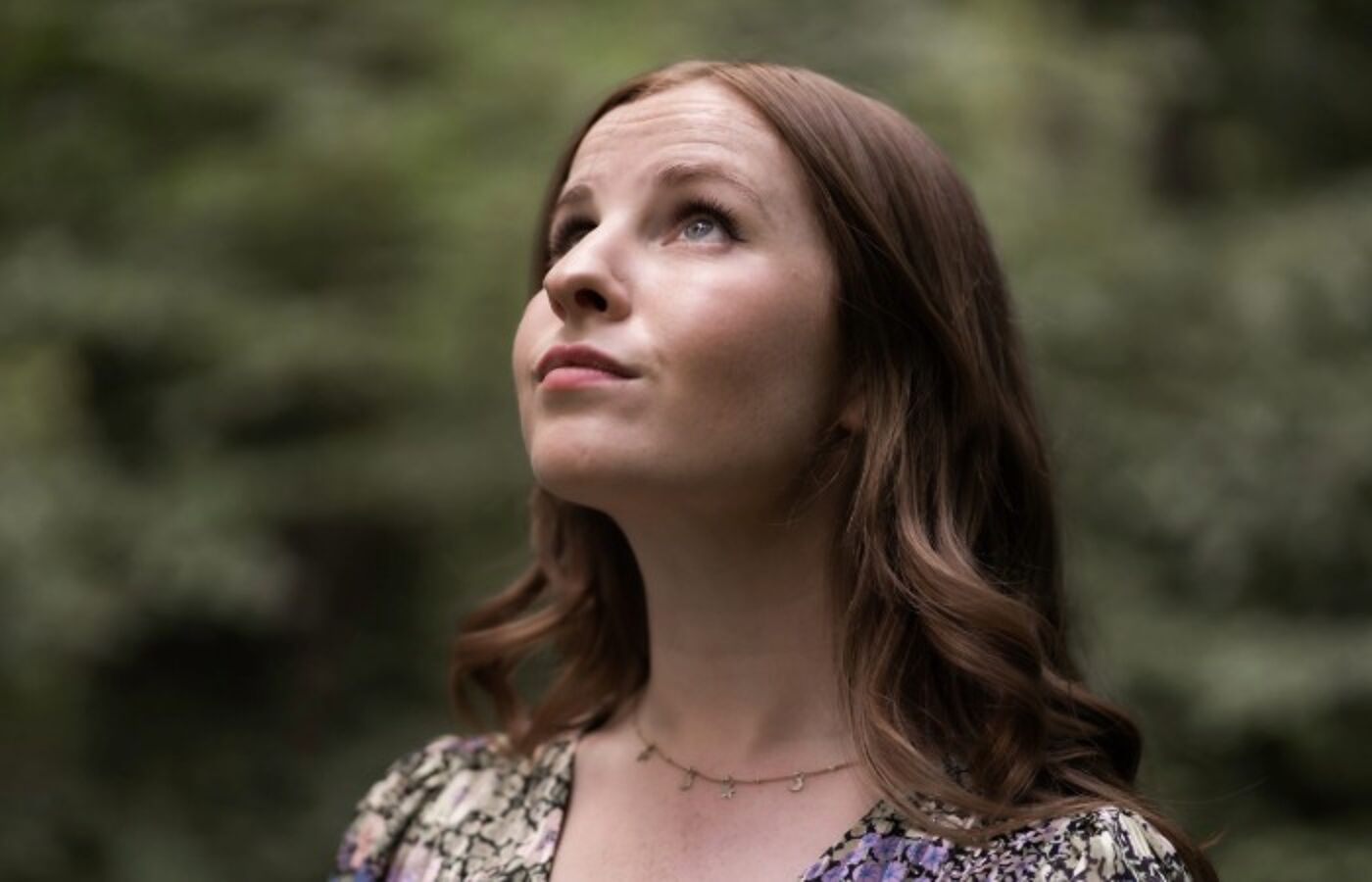 Beth looking up towards the sky, side on to the camera with a blurred background of trees behind her.