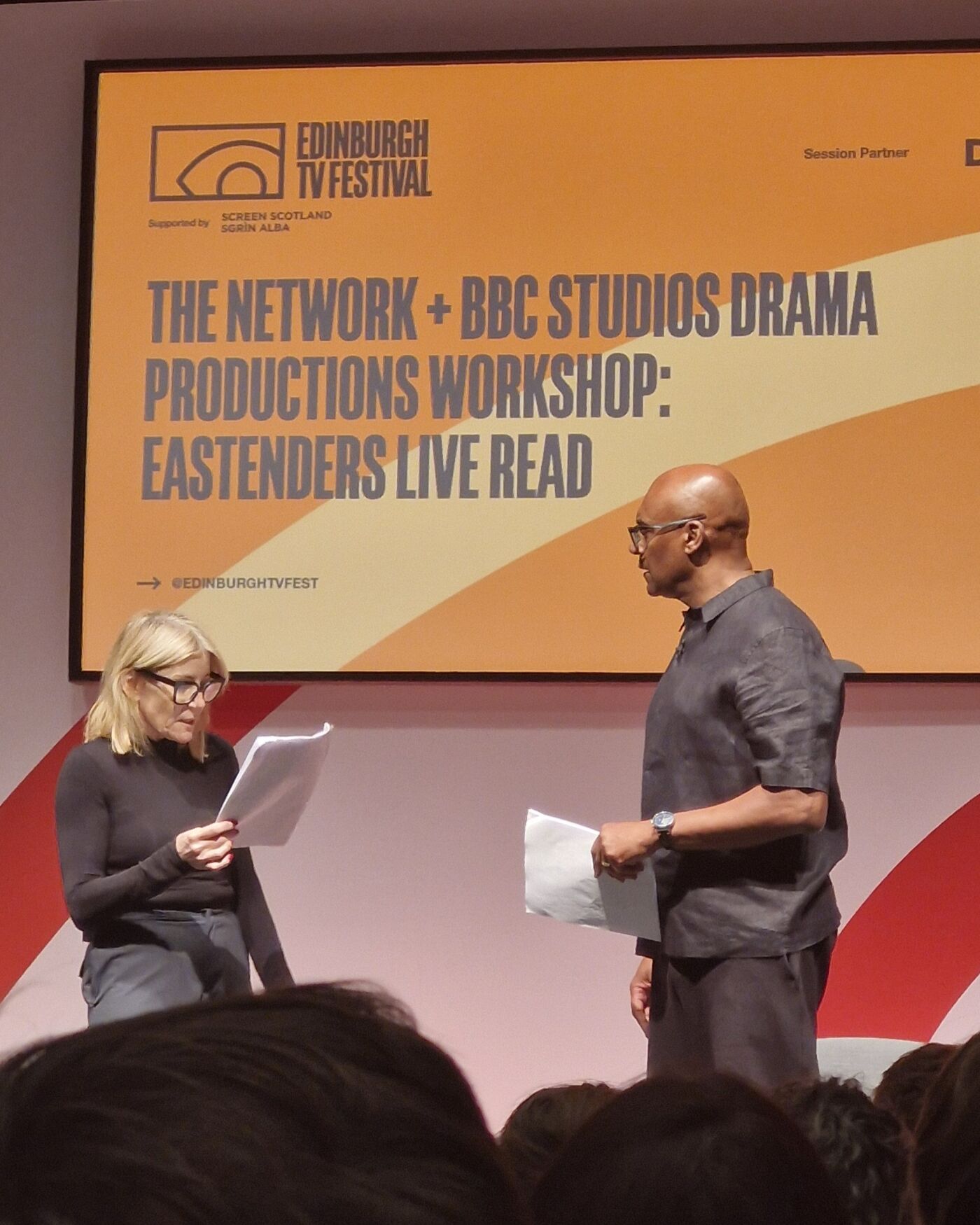 Two people standing in front of a large TV screen reading scripts. The screen reads 'THE NETWROK + BBC STUDIOS DRAMA PRODUCTIONS WORKSHOP: EASTENDERS LIVE READ'