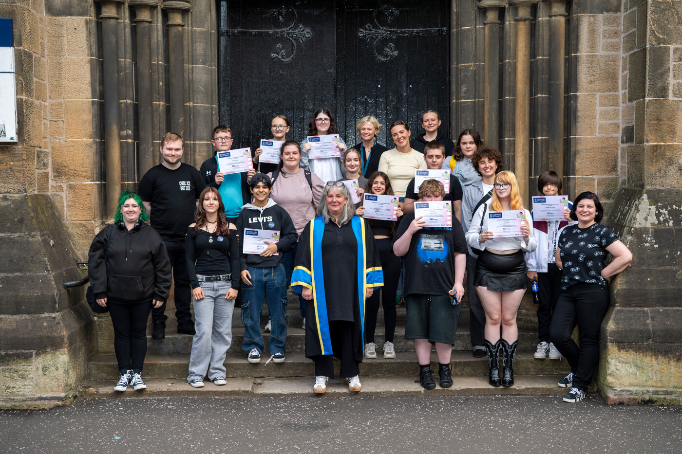A group of people stand on the steps outside Gilmorehill Halls holding their certificates and smiling
