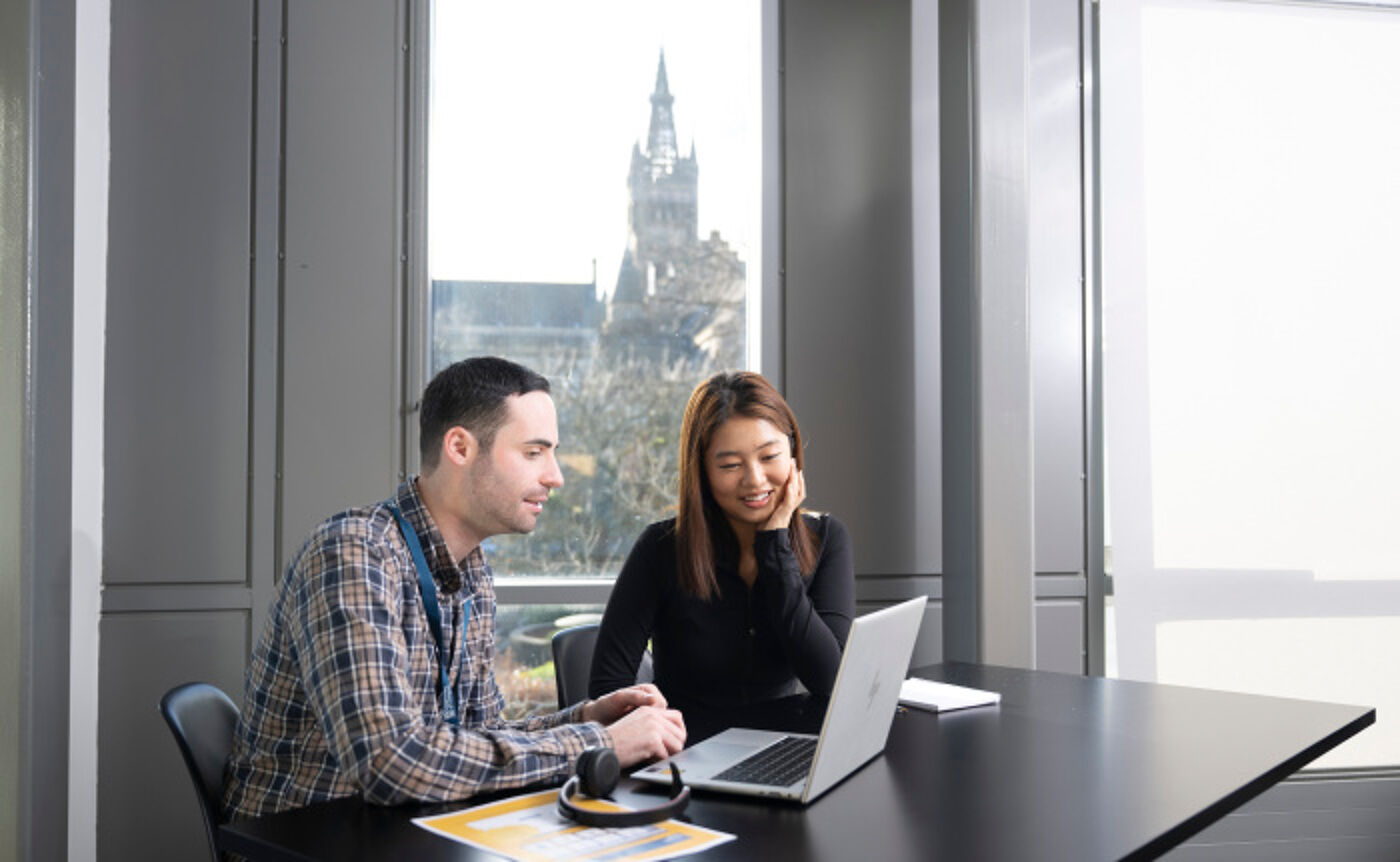 a student and a member of staff sit side by side in a meeting room speaking and looking at a laptop