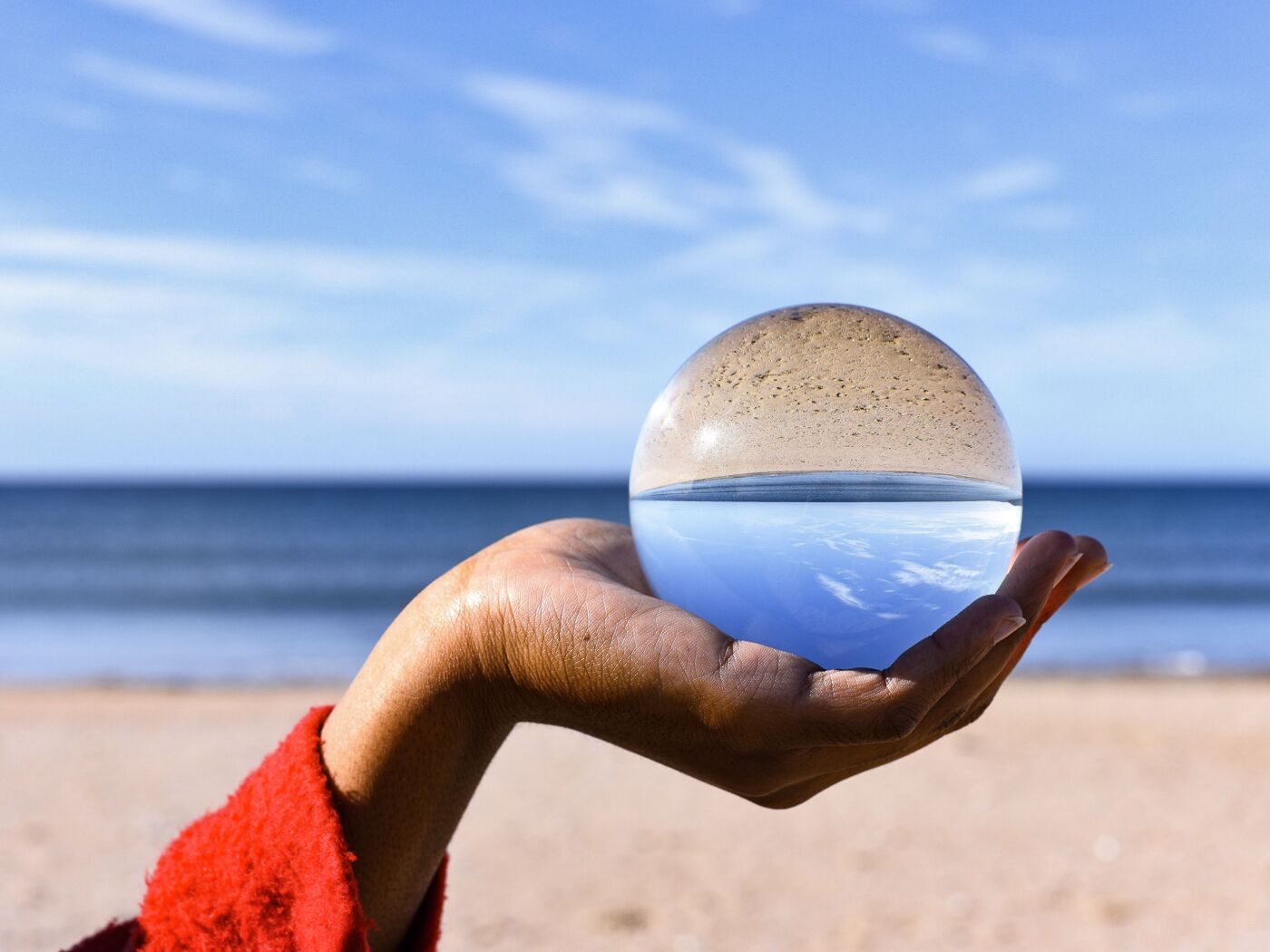 A hand wearing a red sleeve holds a glass sphere that reflects an upside-down view of the beach, sea, and sky on a bright, sunny day.