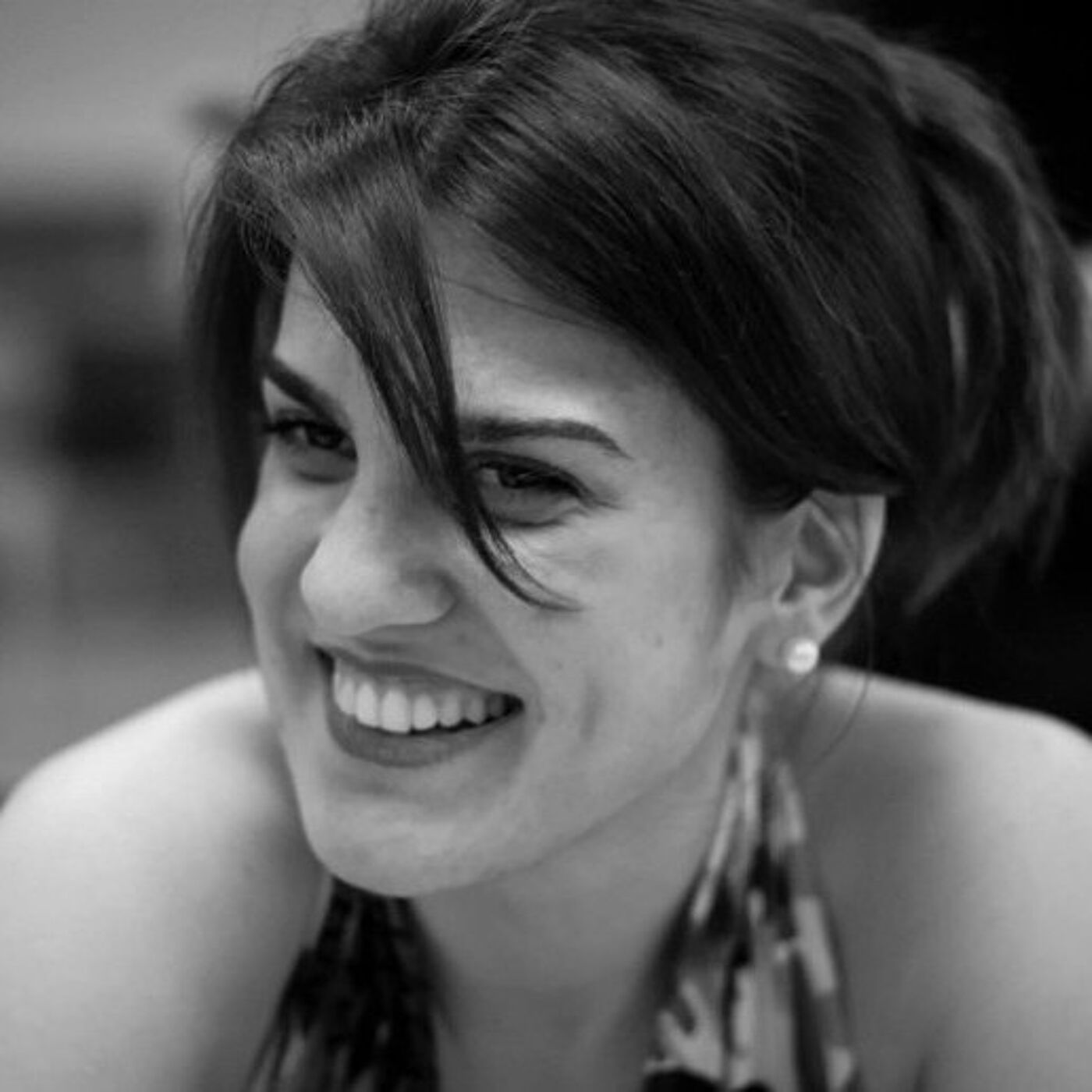 A person with dark, loosely pinned‑up hair is shown in a black‑and‑white photo, wearing a sleeveless patterned top and small stud earrings.