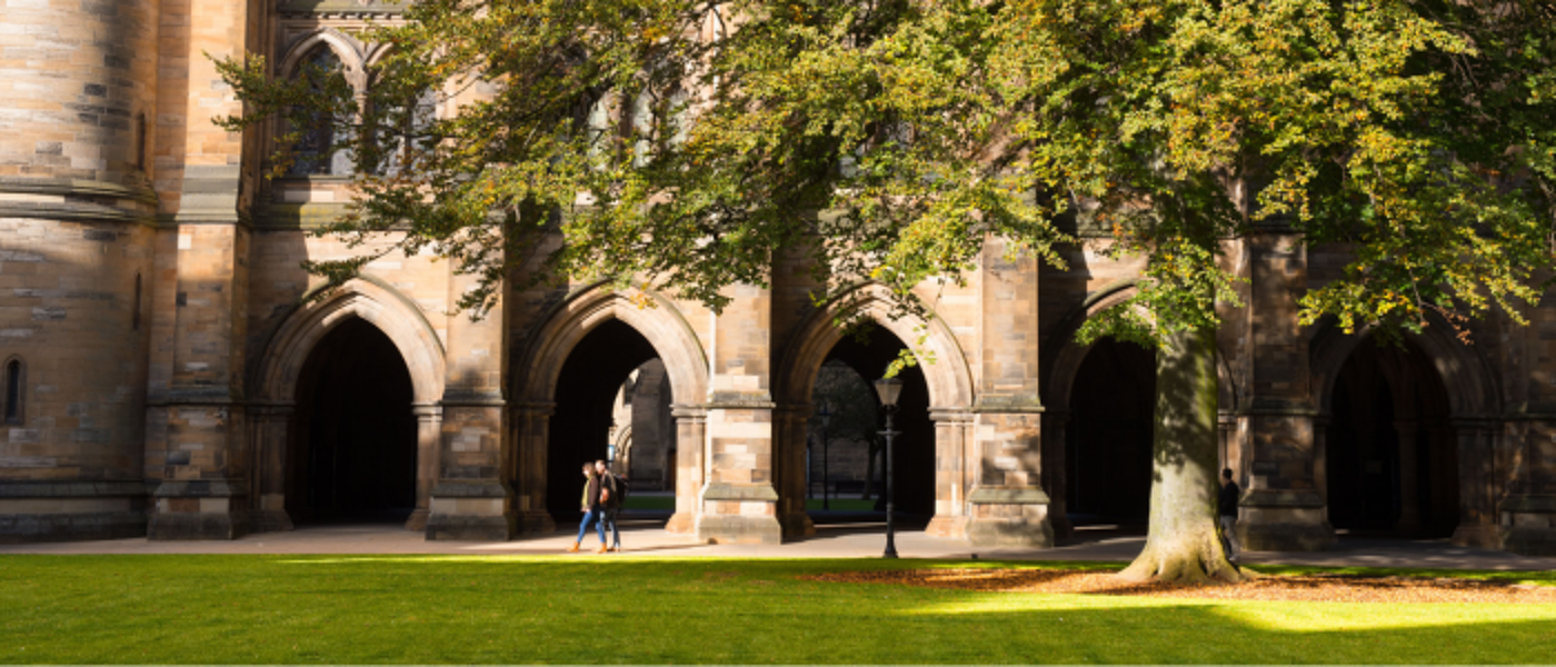 Two people walking through the east quadrangle with the cloisters in the background. Sunlight is shining on the cloisters and tree on the grass.