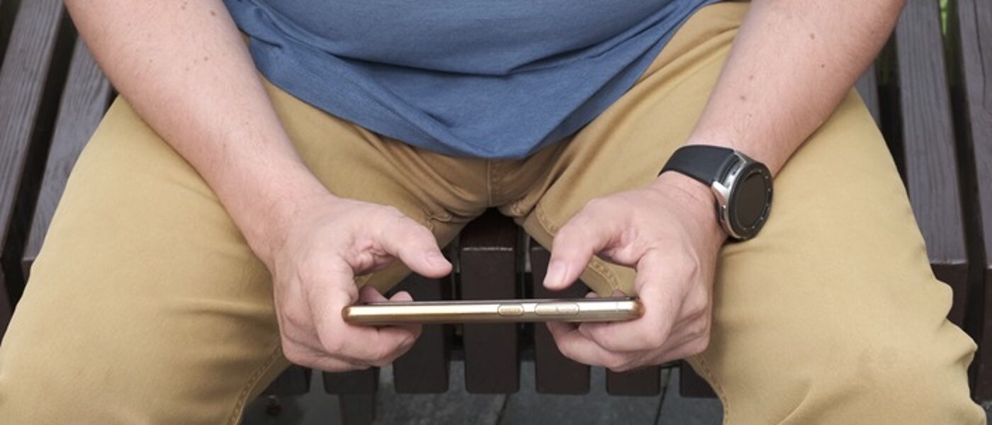 A man wearing a blue tshirt and stone trousers is holding a smart phone in both hands