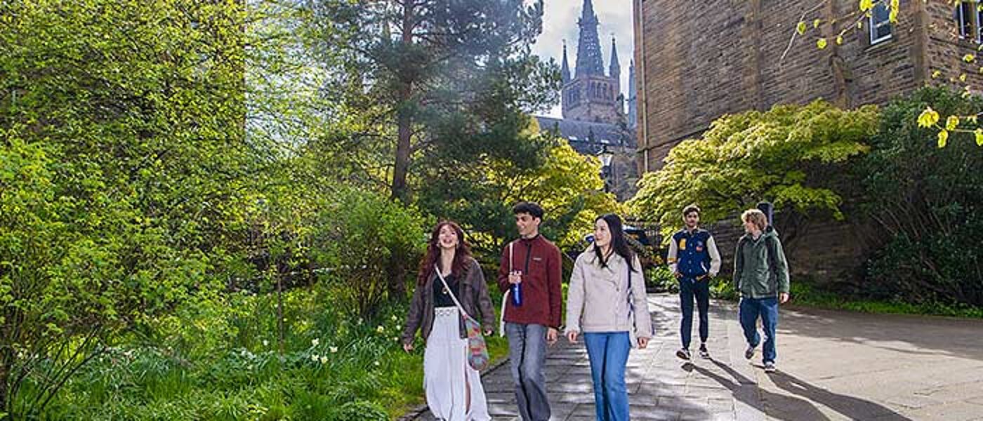 A group of students walking along a path surrounded by green trees and bushes on a sunny day with the historic Gilbert Scott 