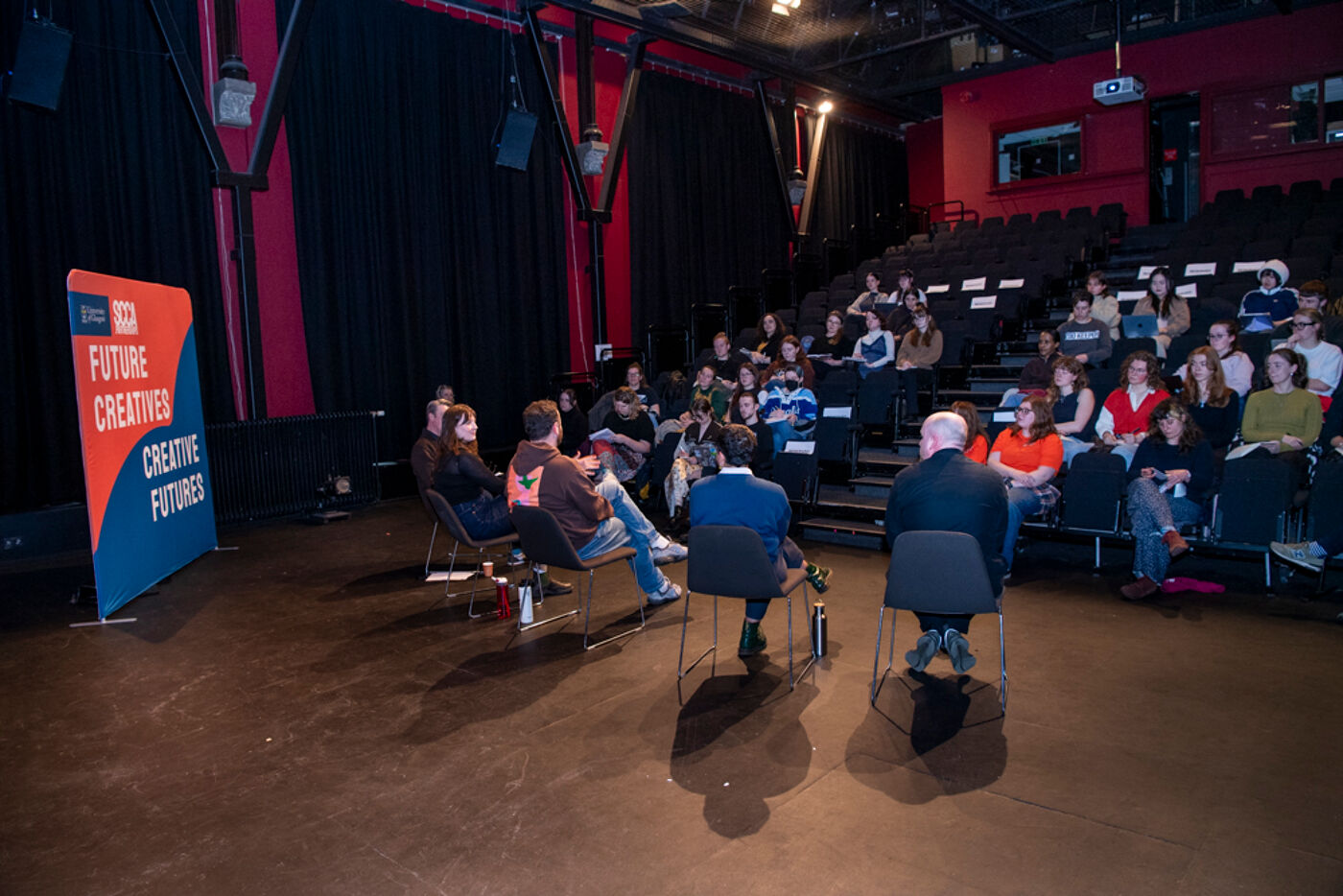 A group of people are seated in a theater-style setting, with a panel in front of them. The banner reads 