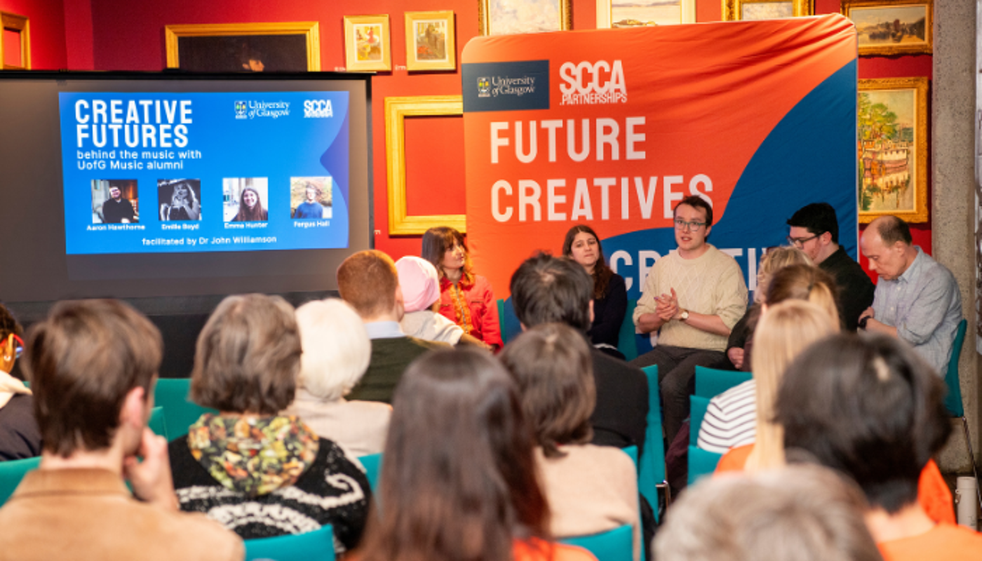 A group of panelists is seated in front of a screen displaying 